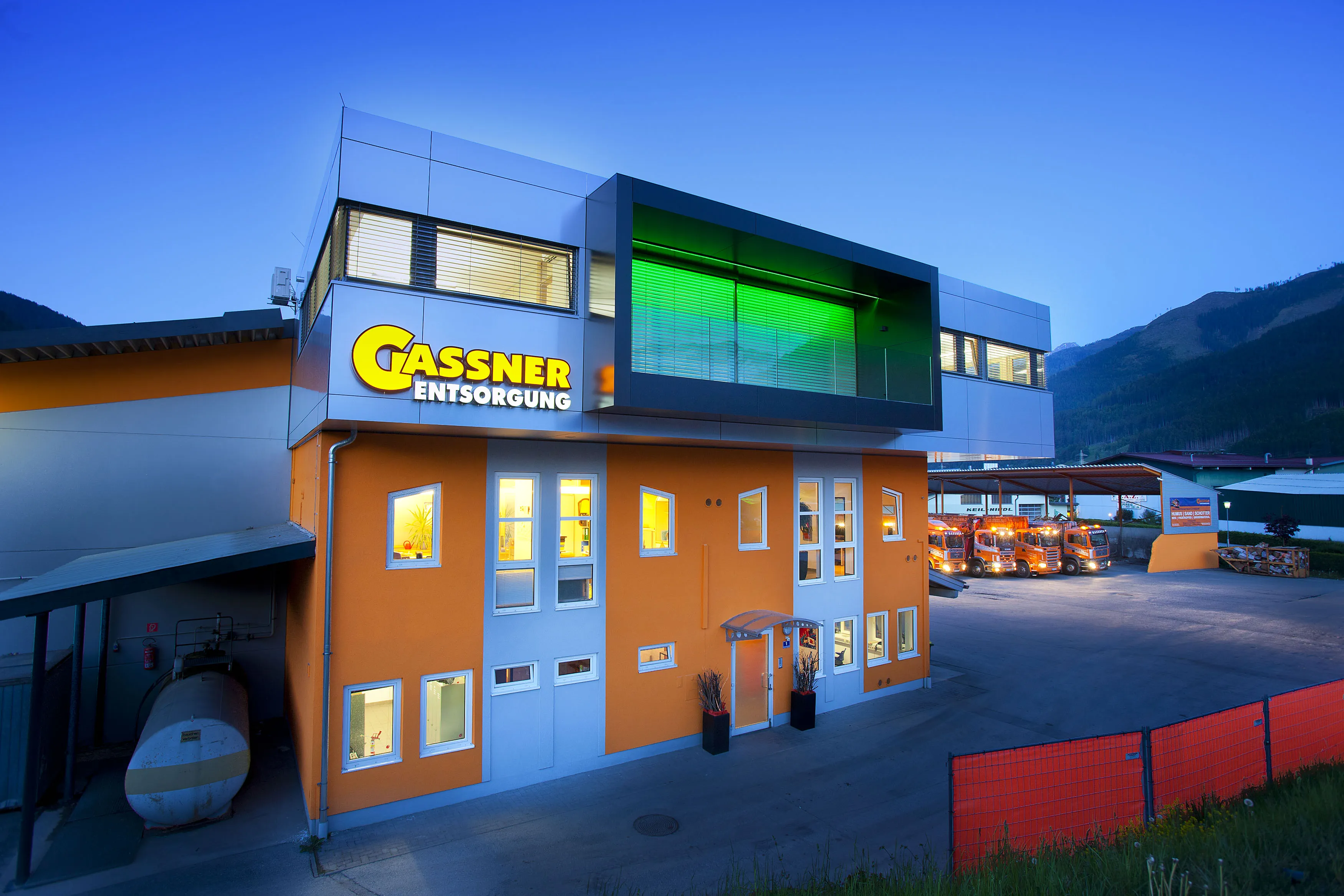 Modern building with large windows, orange facade, and an illuminated sign reading "GASSNER ENTSORGUNG" at dusk. Industrial area visible in the background.