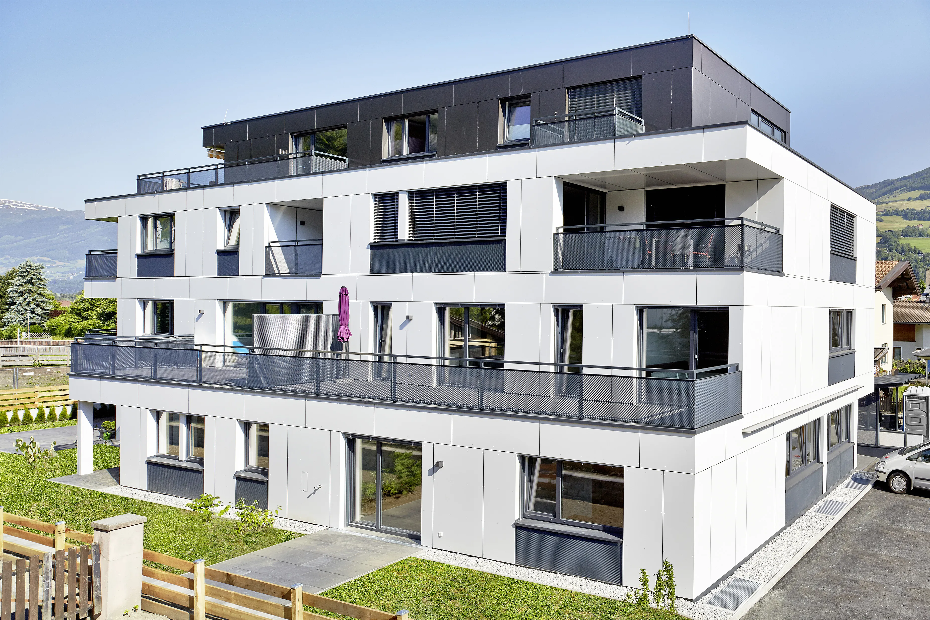 A modern, three-story apartment building with a white and dark gray facade, featuring balconies and surrounded by greenery against a clear sky.