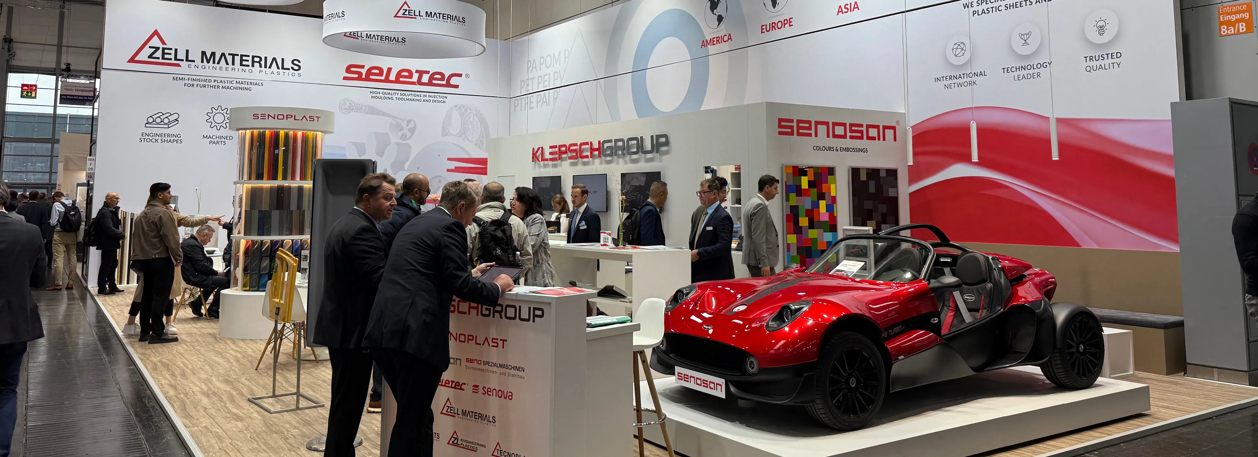 Trade show booth with people gathered around a red open-top sports car and company displays (Zell Materials, Klepsch Group)