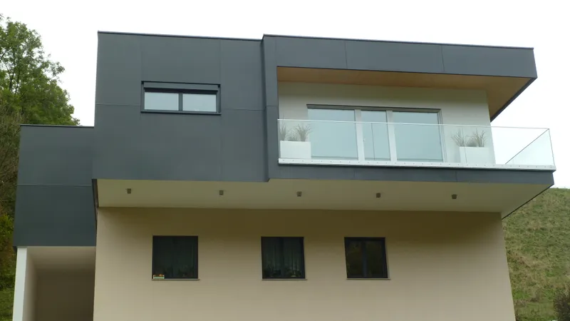 Modern two-story house with minimalist design, featuring a dark upper section, large windows, and glass balcony. Surrounded by greenery.