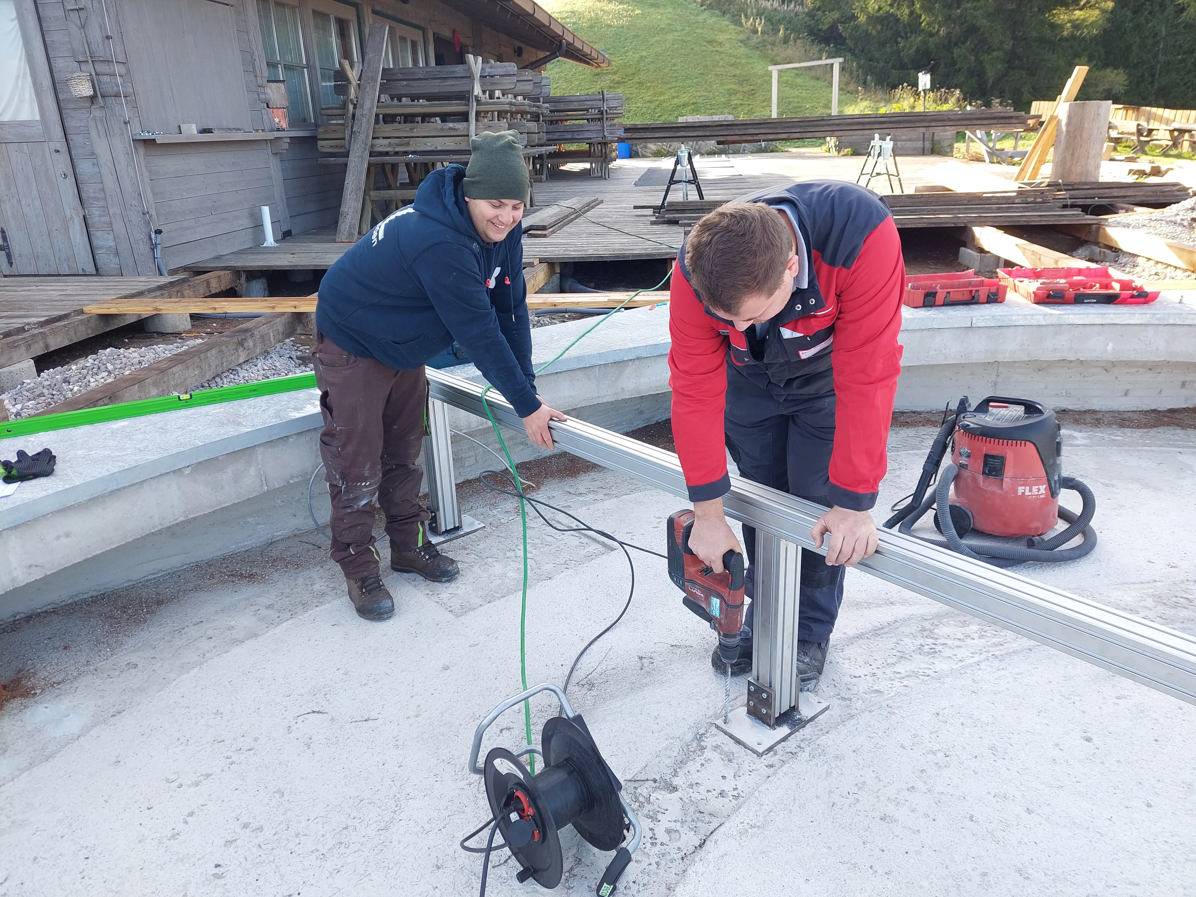 Zwei Mitarbeiter von SENO Spezialmaschinen arbeiten im Freien mit Werkzeugen und montieren Metallkonstruktionen auf einem Betonfundament für die drehbare Terrasse vor einer Almhütte.