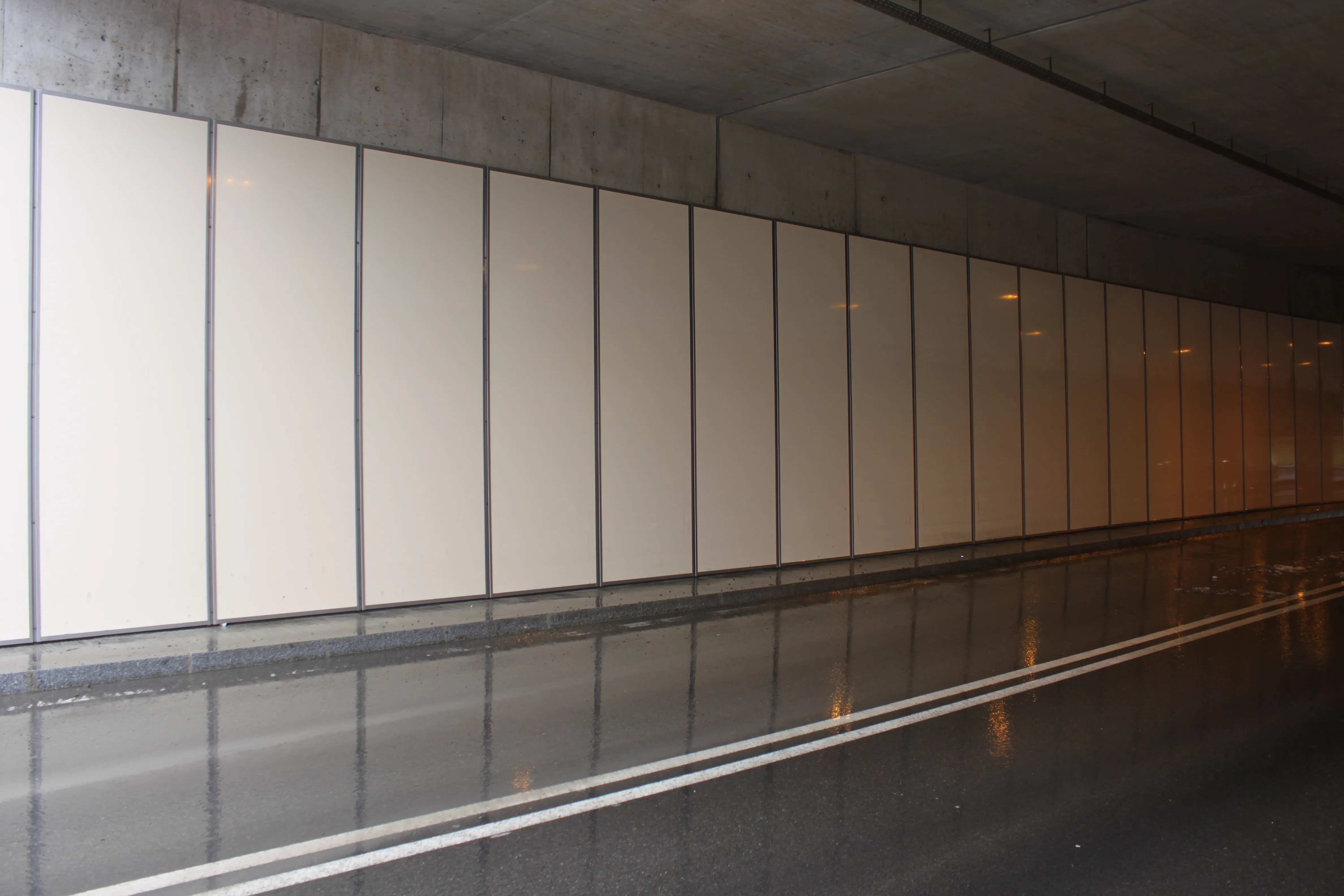 A tunnel interior with smooth cream-colored panel walls and a reflective wet road surface beneath dim lighting.