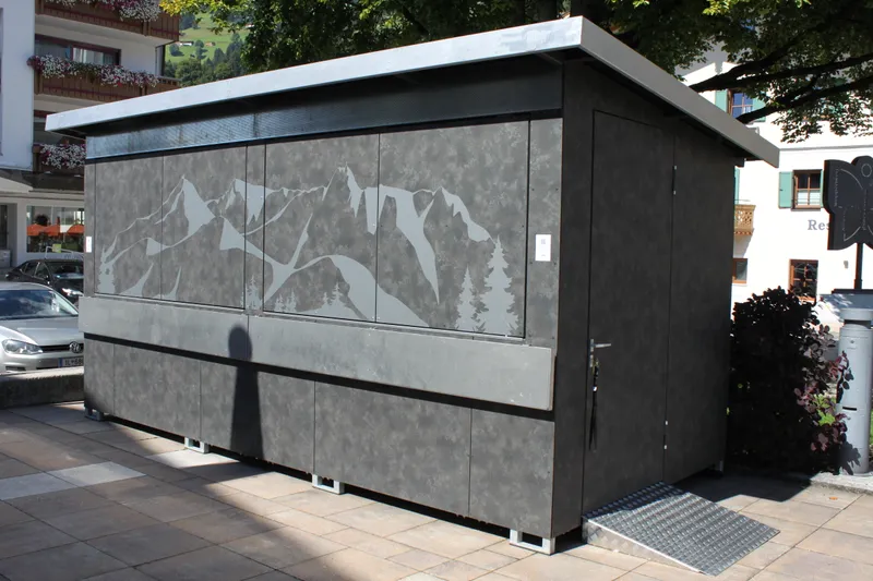 A modern, gray public restroom facility with mountain and tree designs, situated outdoors near buildings and trees.