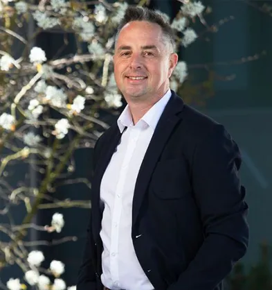 Smiling man in a dark suit and white shirt stands in front of a tree with white blossoms, under soft natural lighting.
