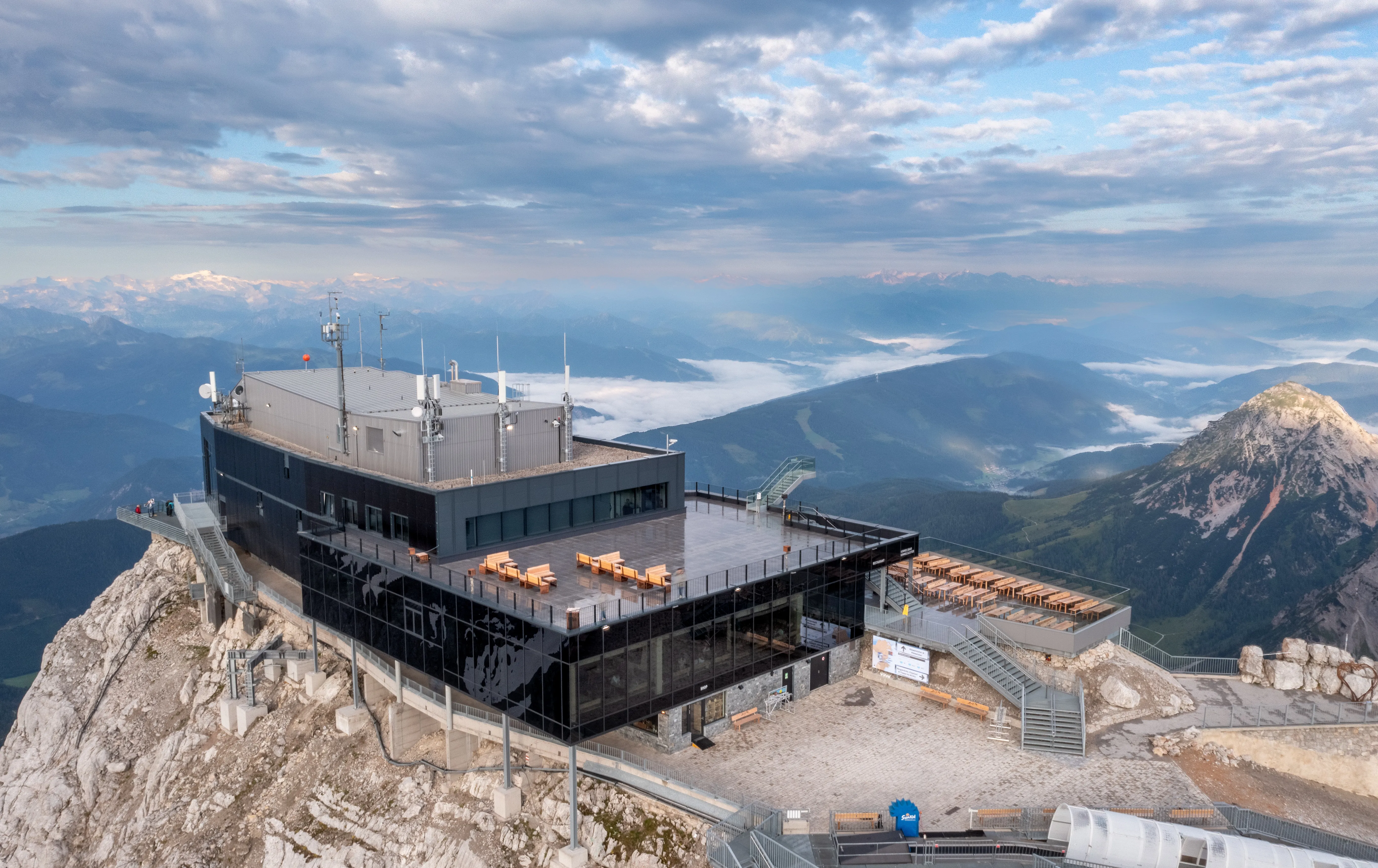 Modern mountain-top building with glass walls, outdoor seating, and scenic views of distant mountains and clouds under a blue sky.