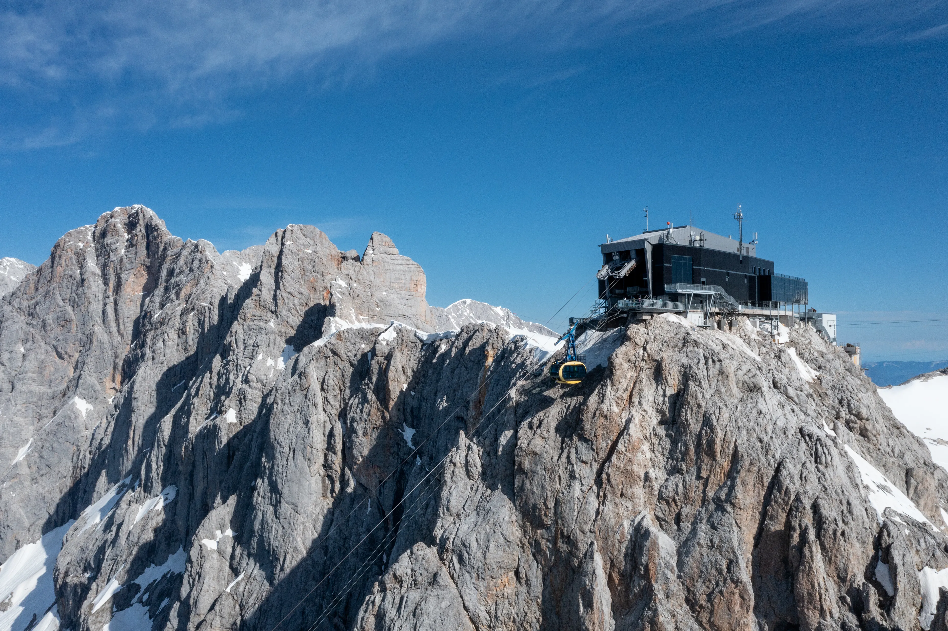 Mountain observatory perched on a rocky peak with snow patches, under a clear blue sky. Rugged terrain and cables visible.