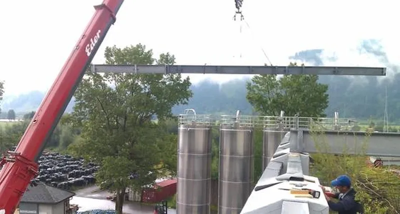 Ein roter Kran hebt einen von SENO Spezialmaschinen gefertigten, großen Metallträger über ein Dach in der Nähe von Industriesilos.