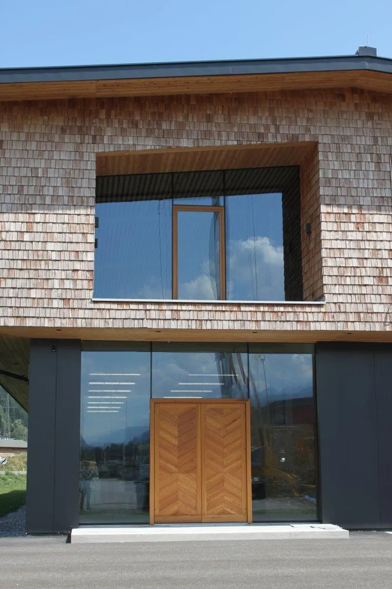 Modern building facade with wooden shingles and large glass windows; wooden double doors at the entrance under a clear blue sky.
