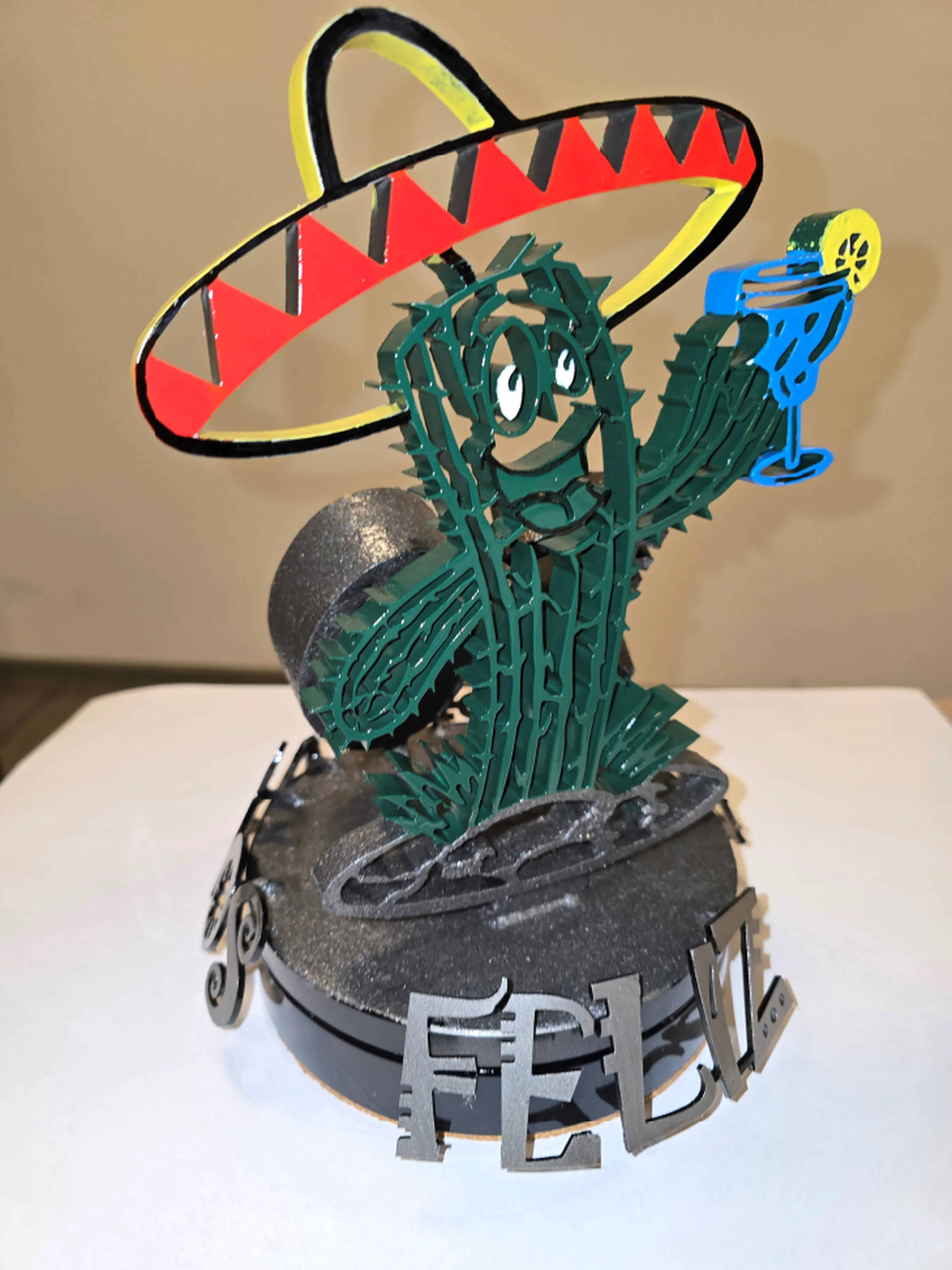 Eine lächelnde Kaktusfigur mit Sombrero hält ein blaues Getränk in der Hand, auf dem Sockel steht „Feliz“.