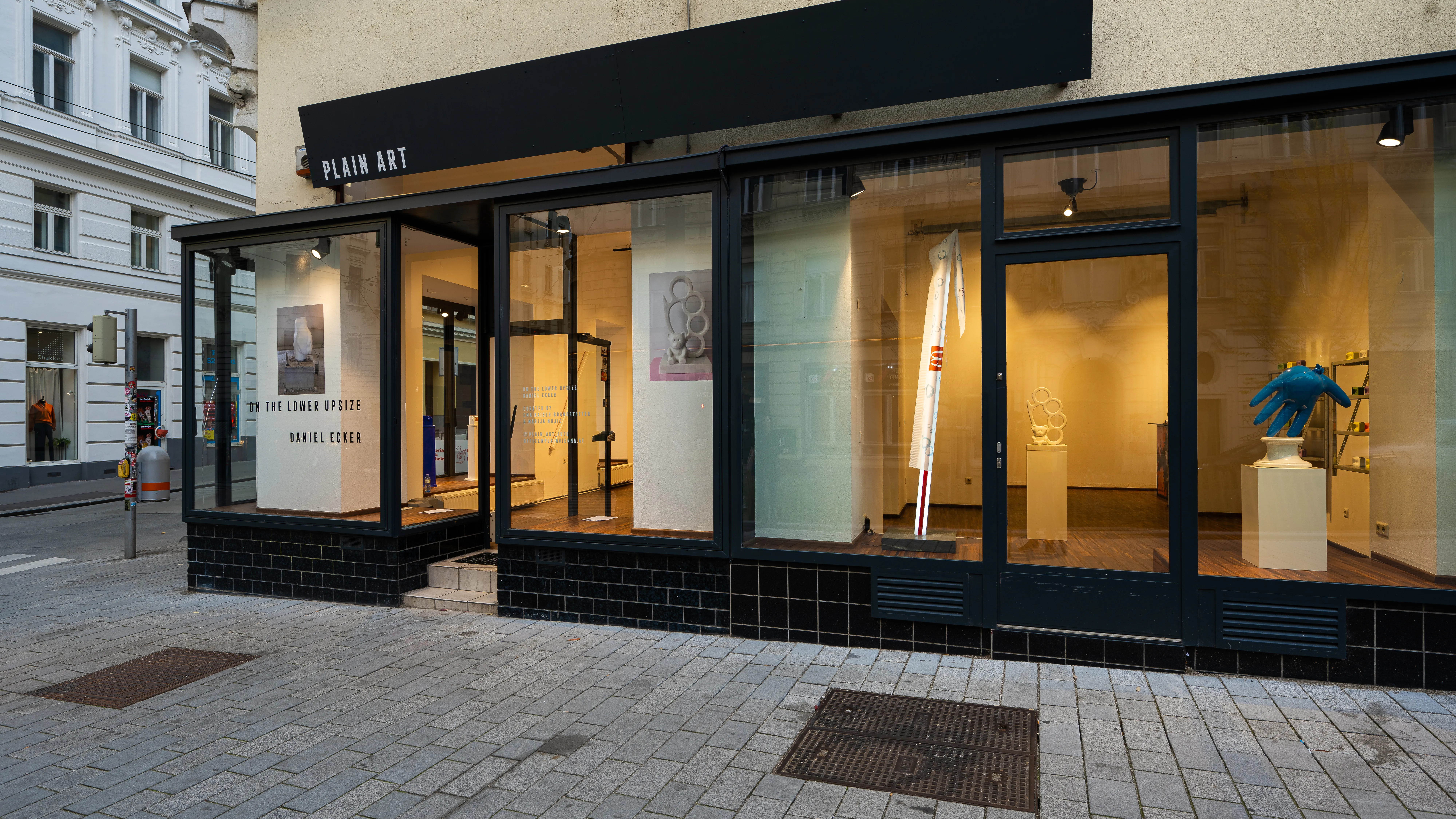 Street view of a modern art gallery with glass windows, displaying sculptures and paintings inside.