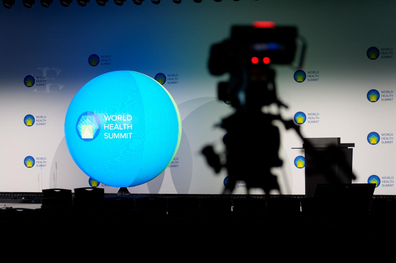 A stage setup with a large blue globe displaying "World Health Summit" text, and a camera silhouette in the foreground.