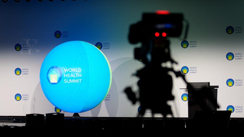 A stage setup with a large blue globe displaying "World Health Summit" text, and a camera silhouette in the foreground.