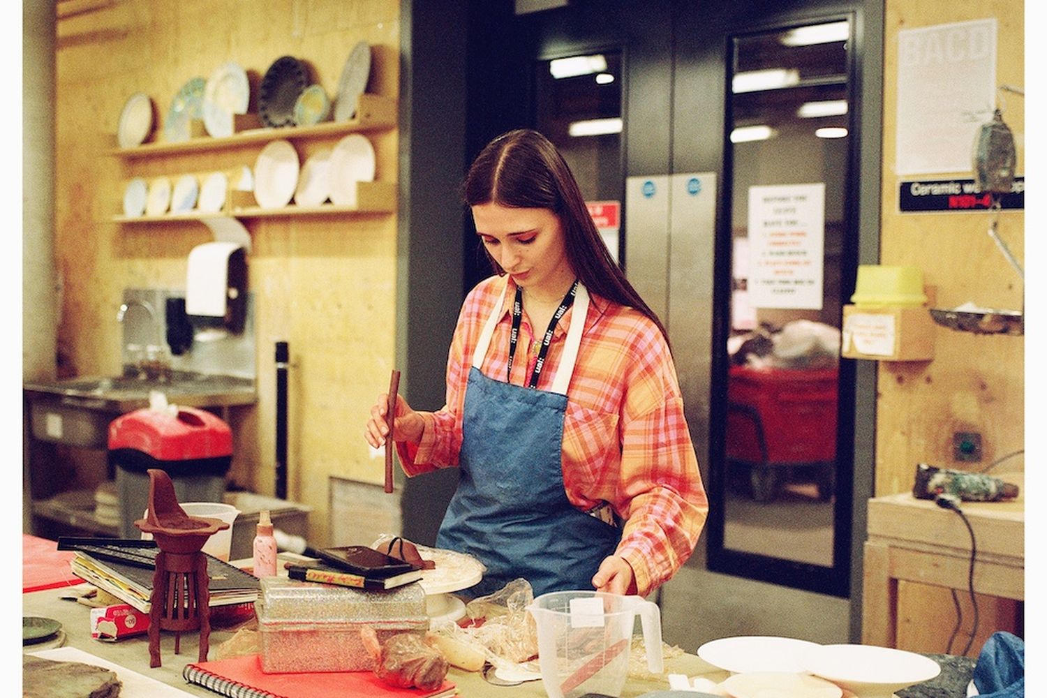 Female artist with long brown hair and orange/pink plaid shirt with a blue apron, weighing clay at a table.