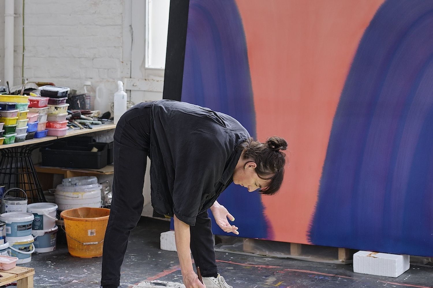 Sara Dare, a professional artist, is in the process of painting a large abstract painting in a studio. She is using a green paintbrush to apply the paint to the canvas. The painting is large and has a vibrant blue and green color scheme. The artist is focused on the task at hand, surrounded by various tools and supplies. [G]