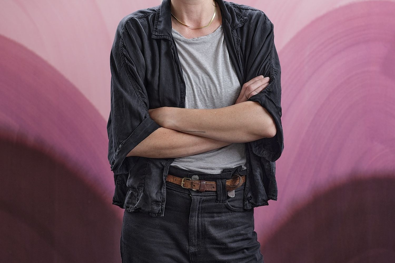 Sara Dare, a young woman with dark hair, stands confidently in front of a vibrant pink and purple abstract painting. She wears a black jacket, a white t-shirt, and black jeans, with a brown belt cinching her waist. Her arms are crossed, and she gazes directly at the camera, creating a sense of connection with the viewer. [G]