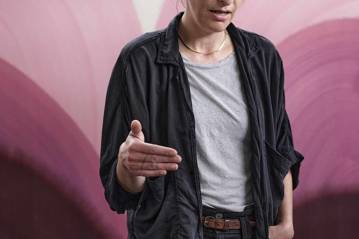 Sara Dare, a woman with short, dark hair, is standing in front of a vibrant pink and purple wall. She is wearing a black jacket, a white t-shirt, and black pants. Her hands are outstretched, suggesting she might be giving a speech or presentation. [G]