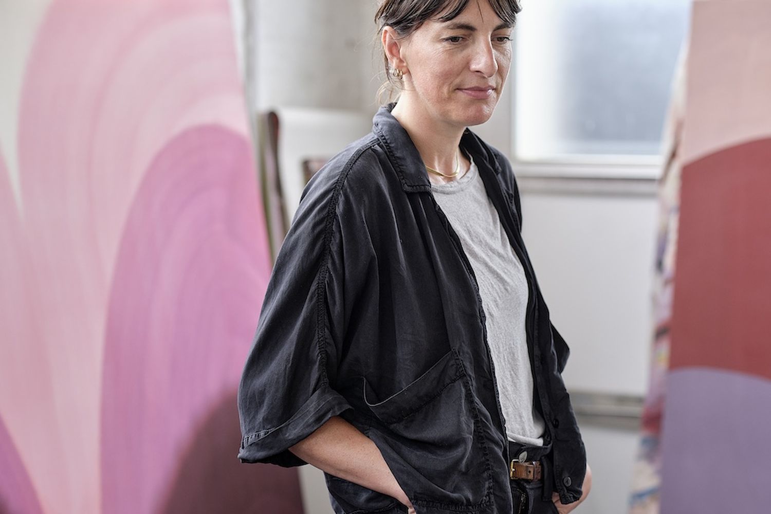 Sara Dare, a woman with short, dark hair, is standing in front of a large, colorful painting. She is wearing a black jacket and a white shirt, and has a brown belt around her waist. She is looking to the side, possibly observing the painting or the surroundings. [G]