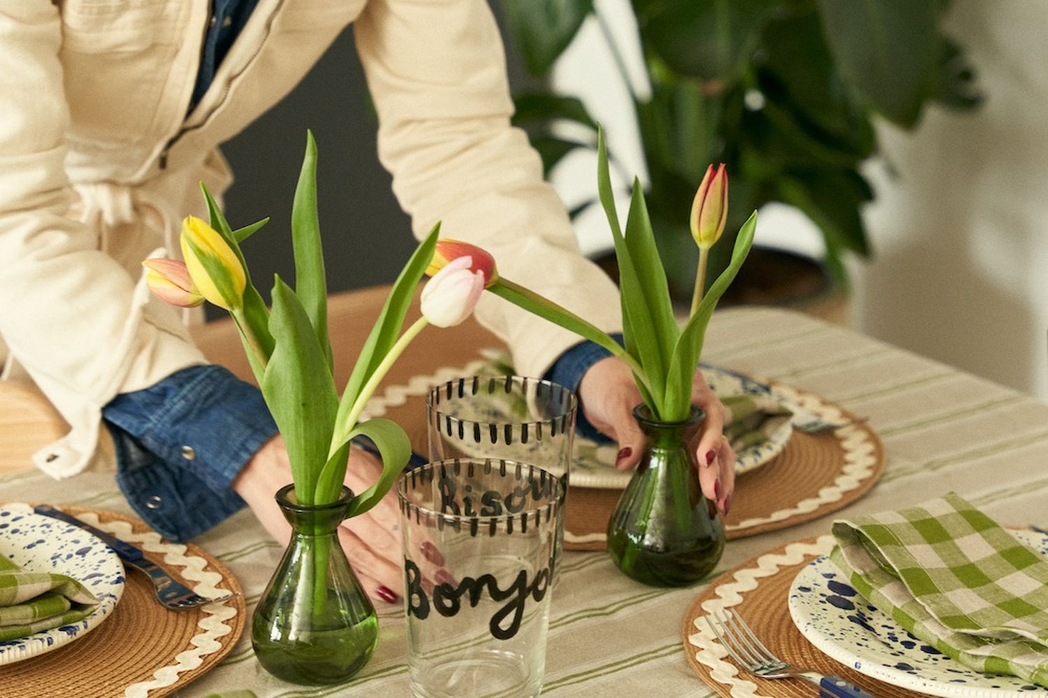 A woman wearing a white boiler suit with a denim shirt under sets green vases with tulips on a dressed table.