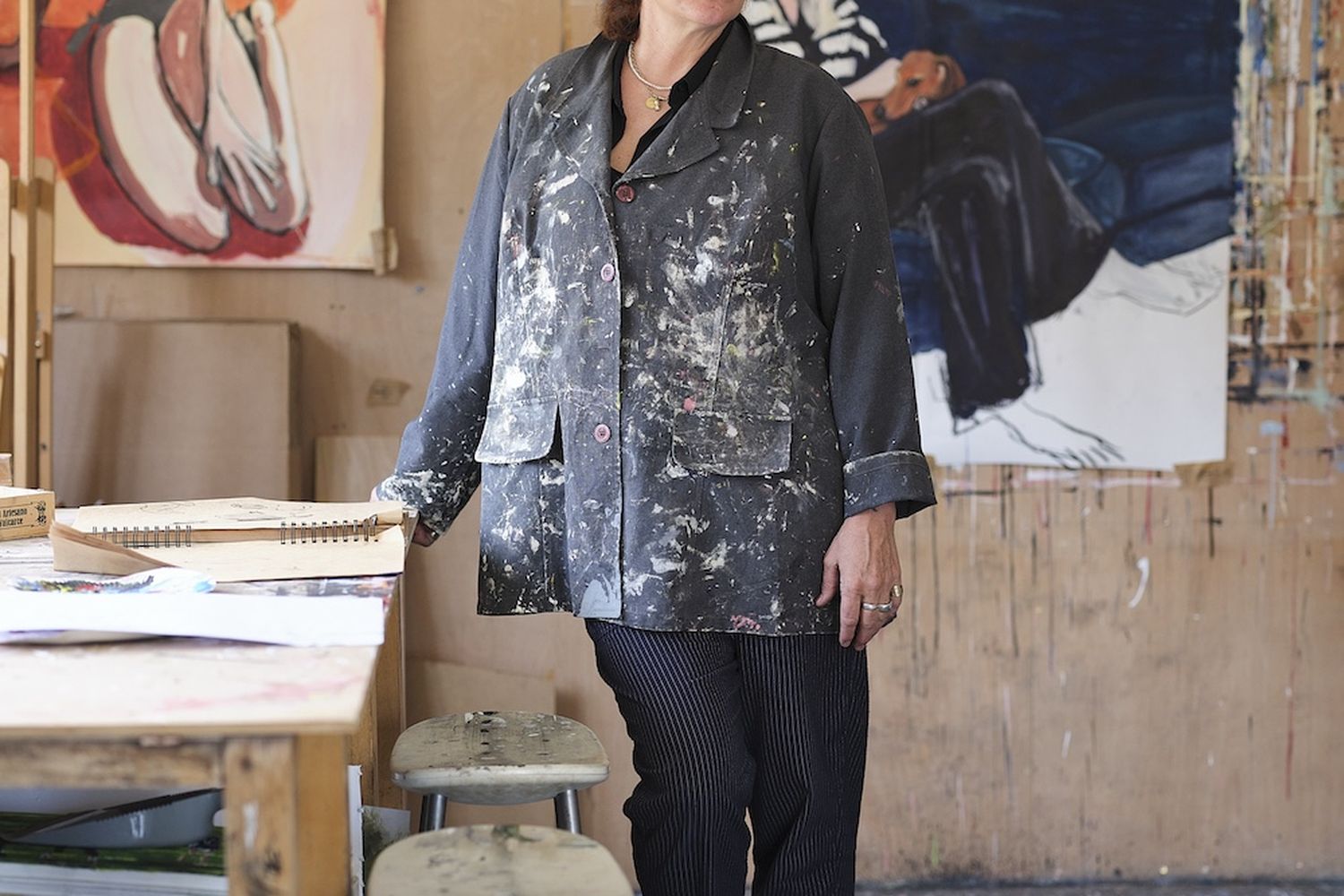 A woman in a blue and white patterned jacket stands in a room with a painting of a woman on the wall. She has glasses and a necklace. There is a stool and a painting on the wall. [G]
