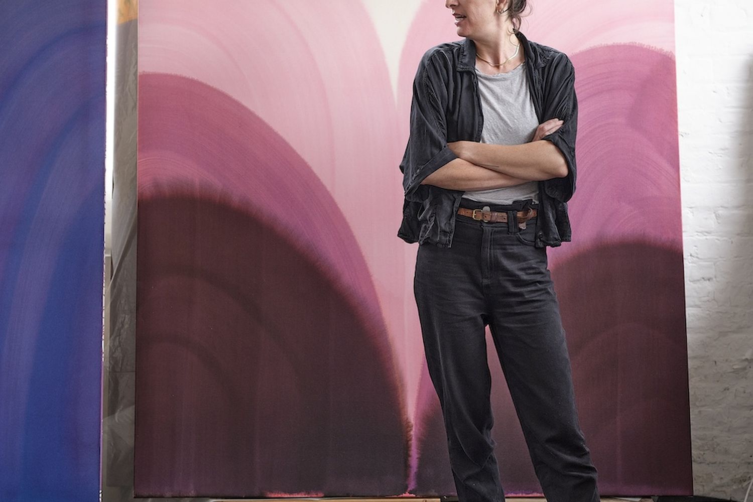 Sara Dolce, a woman with a ponytail, stands confidently in front of a large, abstract painting. She wears a black shirt, dark pants, and white sneakers. The painting, a vibrant mix of pink and purple, is hung on a wooden pallet. The woman's gaze is directed to the left, and her posture suggests she is in a contemplative or artistic state. [G]