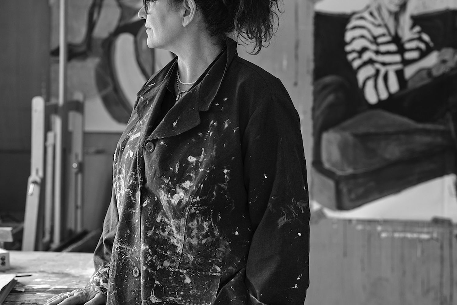 A woman in a black jacket with paint splatters on her sleeves is painting in a studio. [G]