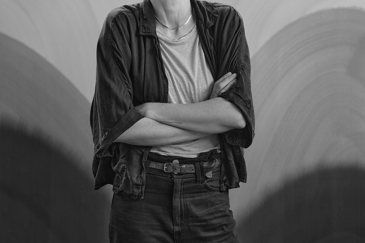 A young man with crossed arms and a wide smile, wearing a black jacket, jeans, and a white shirt, stands confidently against a textured wall. [G]
