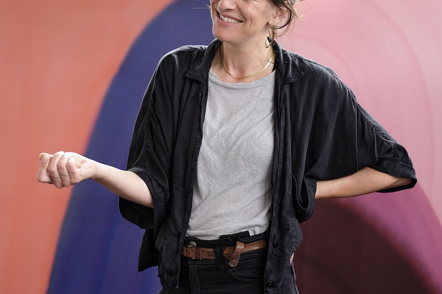 A woman with a bun in her hair, wearing a black jacket and black pants, smiles while standing in front of a colorful painting. [G]