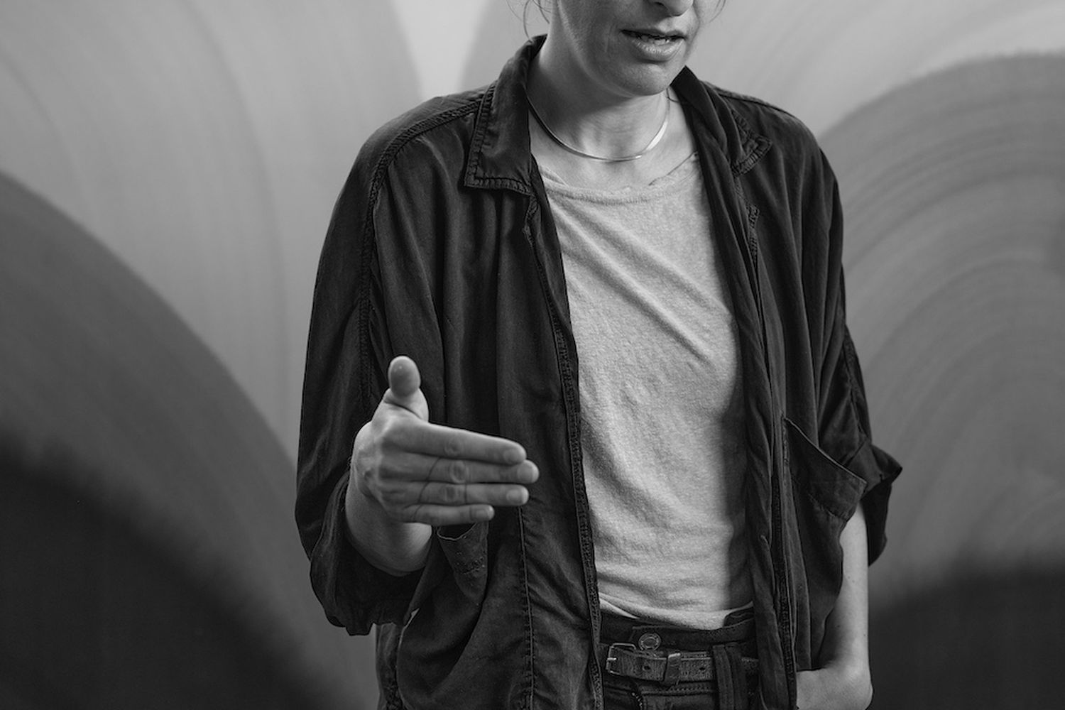 Sara Dare, a woman, stands in front of a wall with circular patterns. She wears a black jacket, jeans, and a white t-shirt. Her hands are raised, and she appears to be speaking or presenting something. [G]