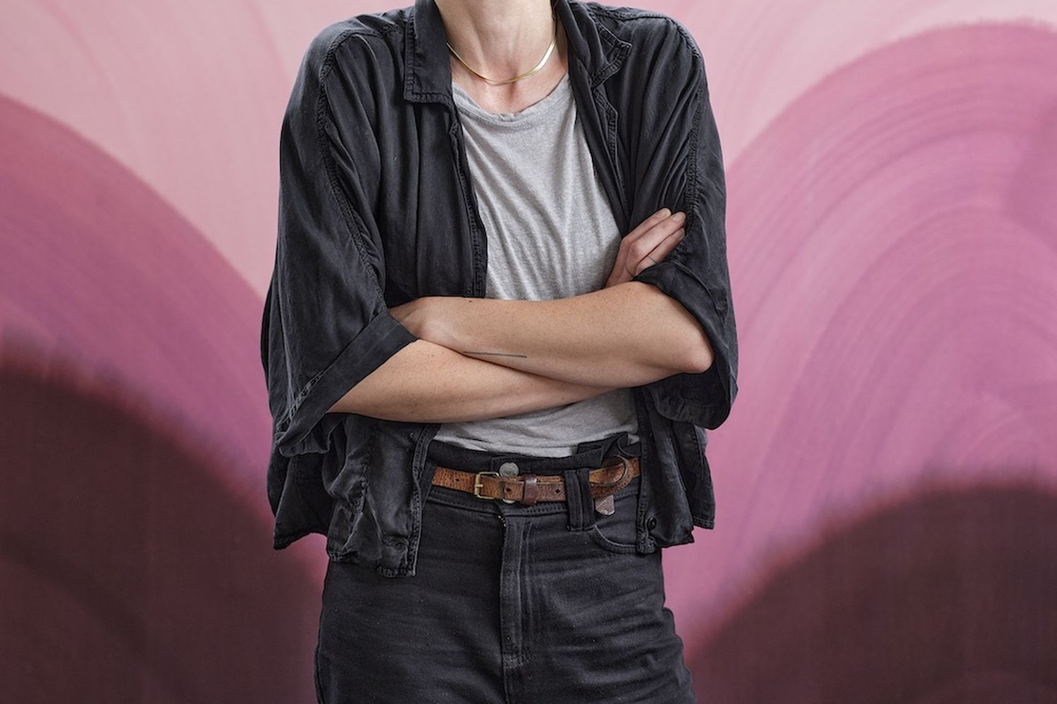 A young man with a wide smile, wearing a black jacket and black pants, stands confidently in front of a vibrant pink and purple wall. [G]