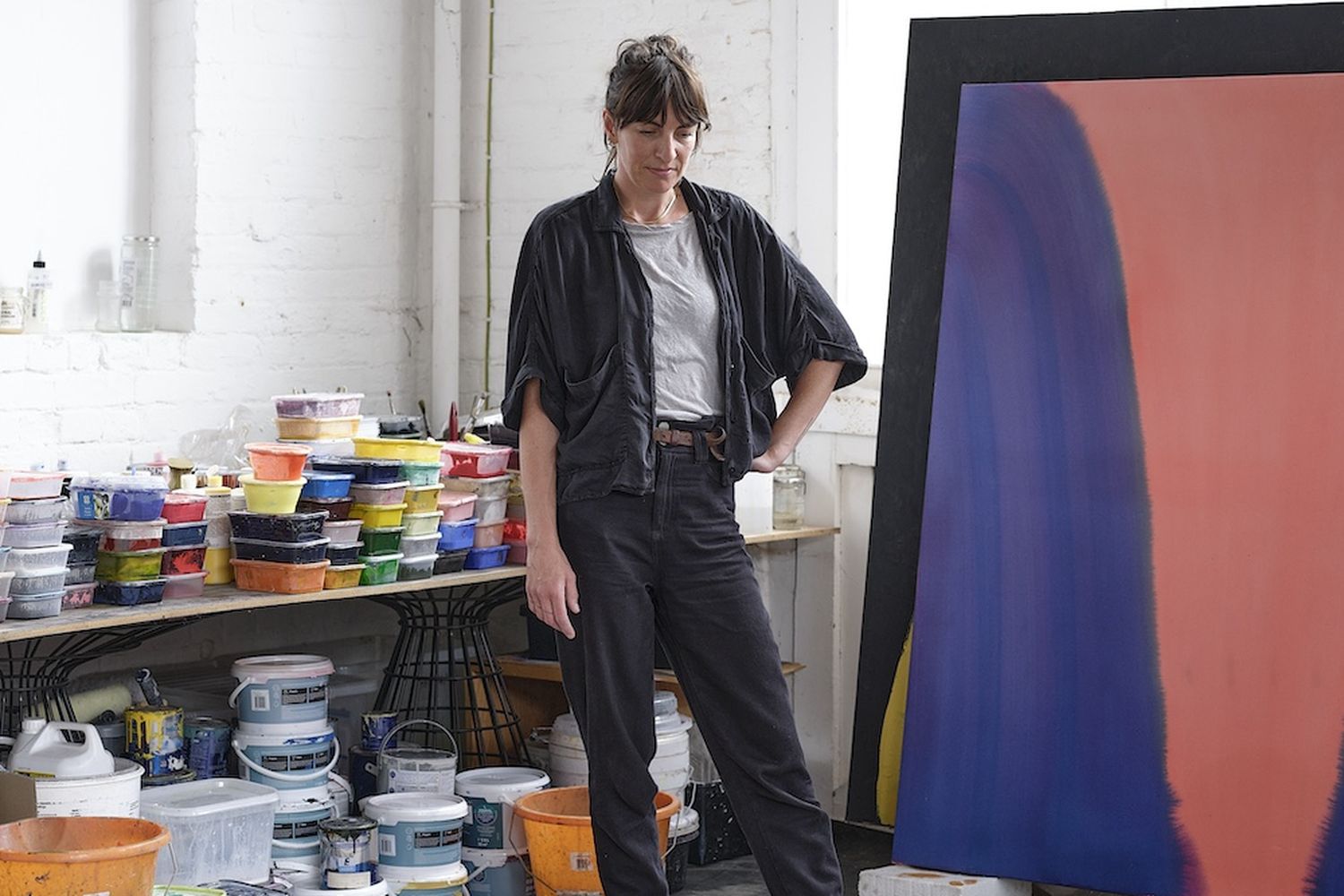 Sara Dare, a woman in a black overcoat, poses in an artist's studio. The studio is filled with various art supplies and a large abstract painting. [G]