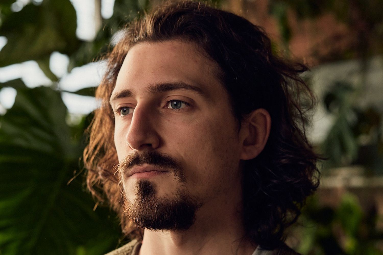 A man with long, curly hair and a beard, wearing a sweater, is looking off to the side in a room filled with green plants. [G]