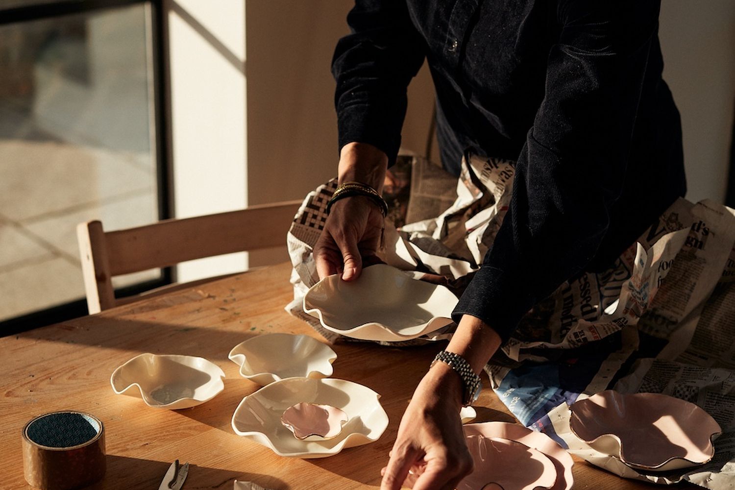 A woman is crafting pottery at a wooden table. She is using scissors and a paintbrush to shape and decorate the pottery. The table has a variety of pottery pieces, including a clay pot and a ceramic bowl. The woman is wearing a denim jacket and a watch. The background shows a window with a view of the outside. [G]