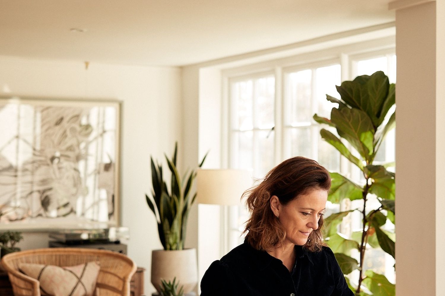 A woman is sitting at a desk, working on a piece of paper. She is wearing a black shirt and has brown hair. The desk has a lamp and a plant on it. The room has a window and a plant is placed near the window. [G]