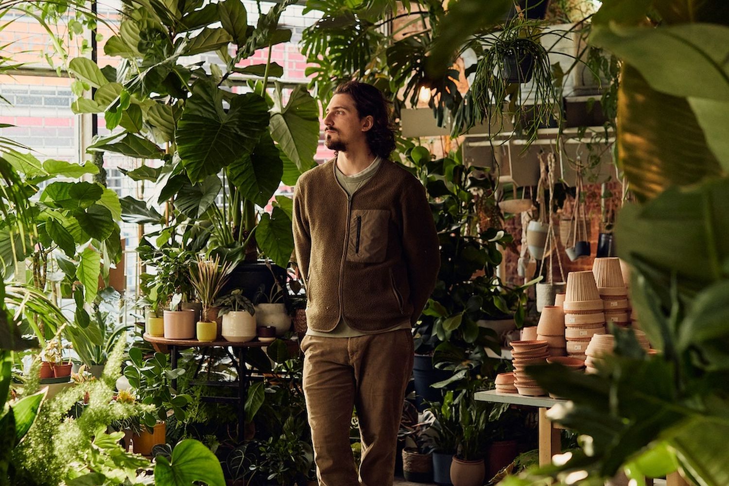 A man stands in a greenhouse filled with various plants, admiring the vibrant greenery. [G]