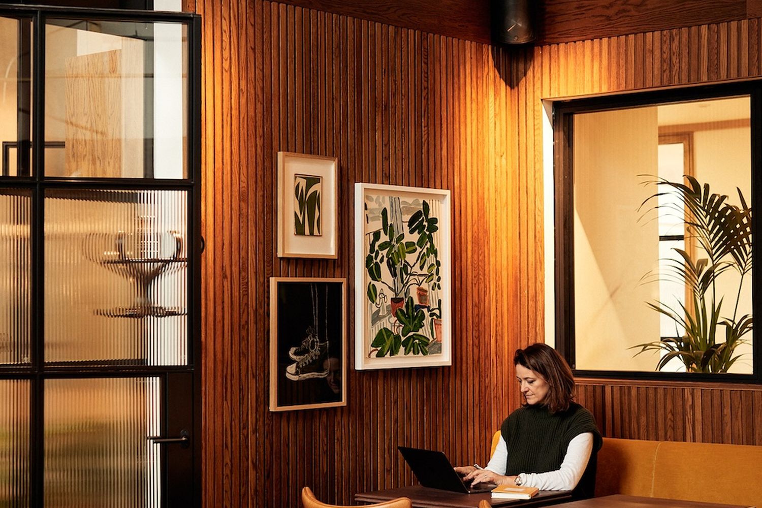 A woman is working on her laptop in a cozy, modern cafe. The cafe has wooden walls and a large window that lets in natural light. There are several paintings on the wall, and a plant is placed near the window. The woman is seated on a brown leather chair, and there are several other chairs around her. [G]