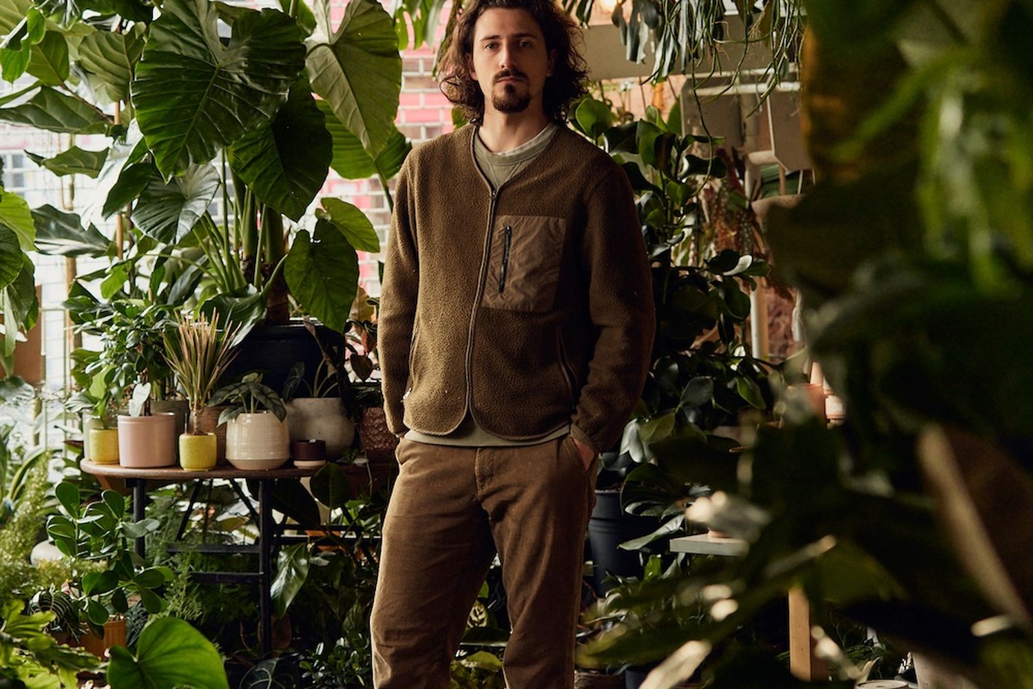 In this image, actor Chris Hemingway is captured in a moment of tranquility amidst a lush greenhouse. He stands confidently, his hands resting on his hips, gazing off to the side with a serious expression. His attire is casual yet stylish, with a brown sweatshirt and matching pants that blend seamlessly into the greenery of the greenhouse. The greenhouse itself is a vibrant haven of life, filled with a variety of plants and flowers that add a touch of nature's beauty to the scene. The image is a testament to Hemingway's ability to capture the essence of his character in a moment of quiet introspection. [G]