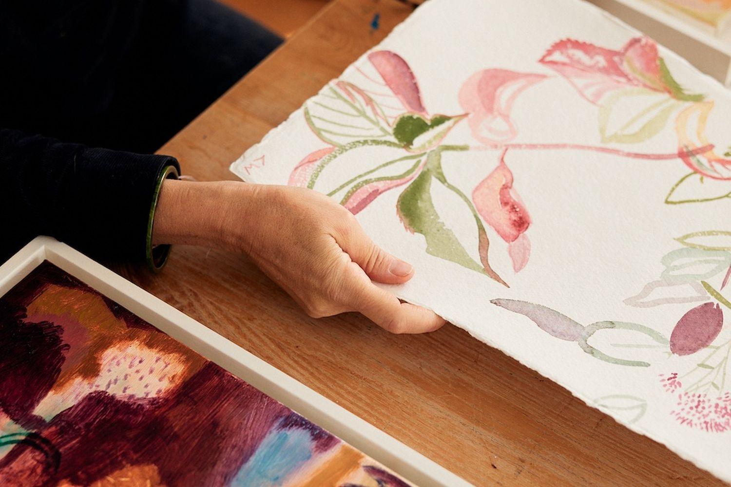 A hand is reaching for a piece of paper with a vibrant floral design, while another piece of paper is placed on a wooden table. The scene suggests a workspace where someone is engaged in art or design. [G]