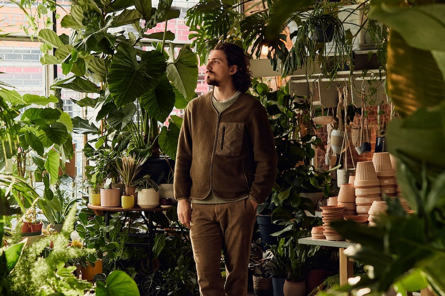 A man with a beard and goatee, wearing a brown jacket and brown pants, stands in a greenhouse filled with various plants and pots. [G]