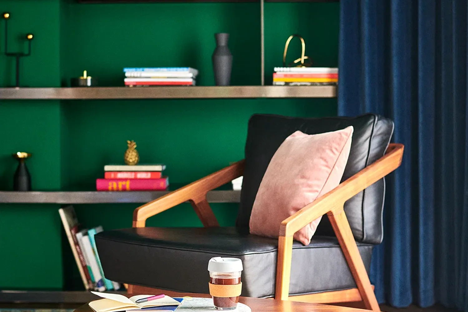 The image features a cozy living room with a black leather chair and a wooden coffee table. The chair is adorned with a pink pillow and a wooden coaster. The coffee table has a cup of coffee on it. The room is decorated with a green wall, a blue curtain, and various objects such as a vase, a candle, and a vase with a flower. The arrangement of objects creates a warm and inviting atmosphere. [G]