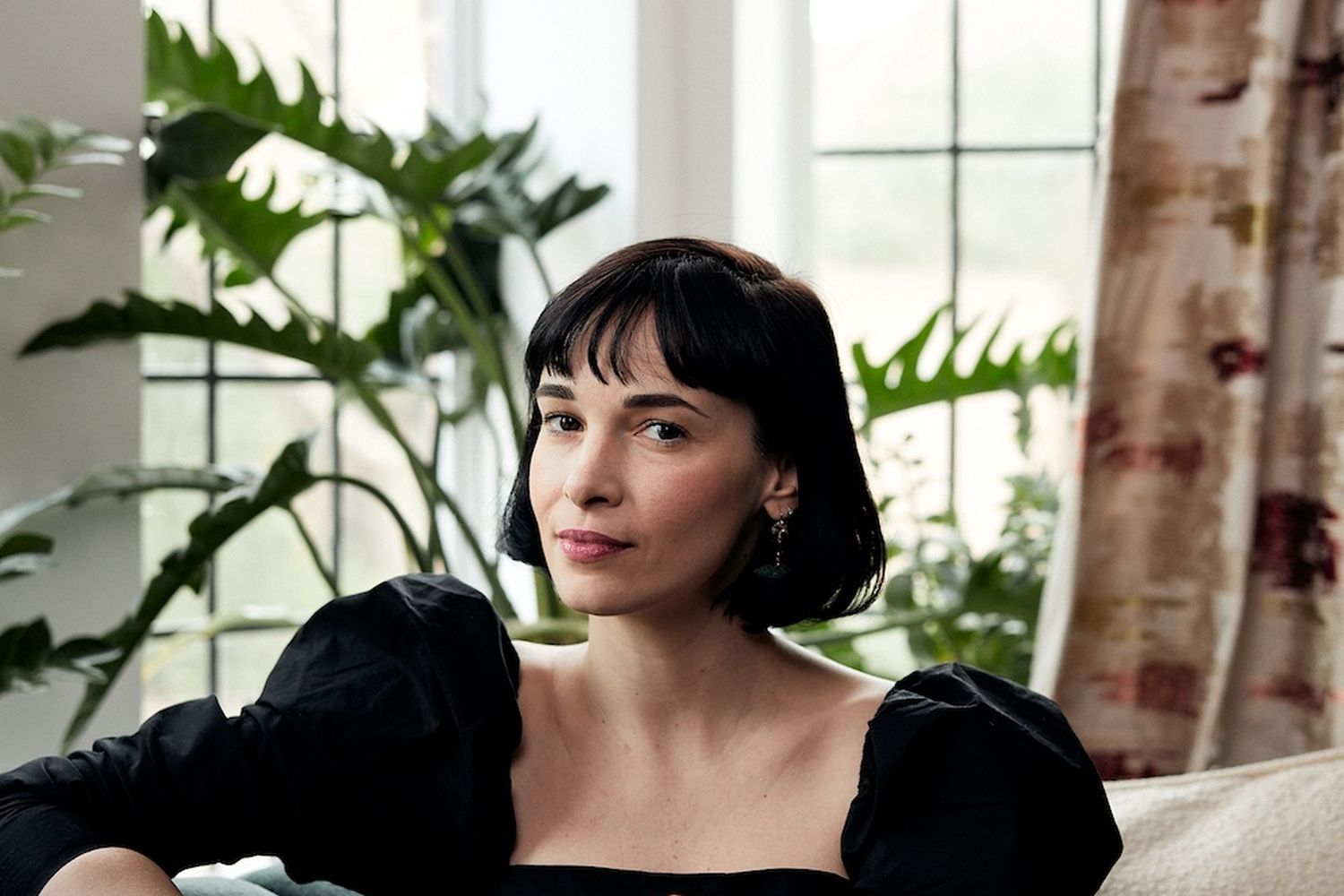 A woman is looking at the camera while sitting on a sofa in front of a window.