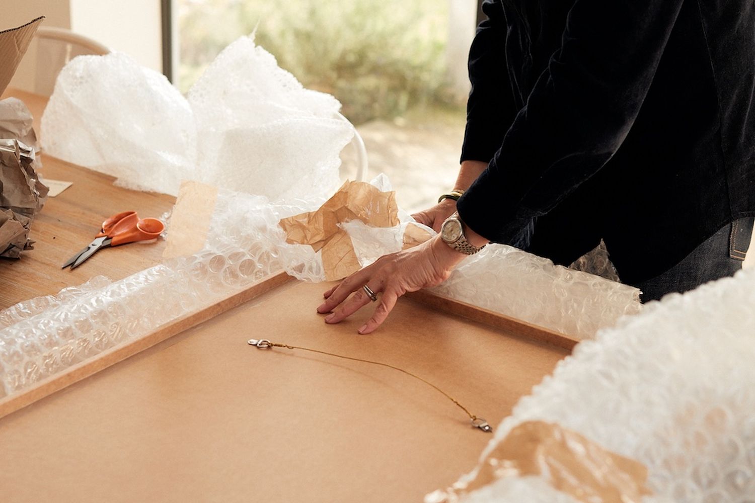 A person is working on a cardboard box, with a pair of scissors nearby. The box is on a table, and there are other objects like a bag and a box. [G]