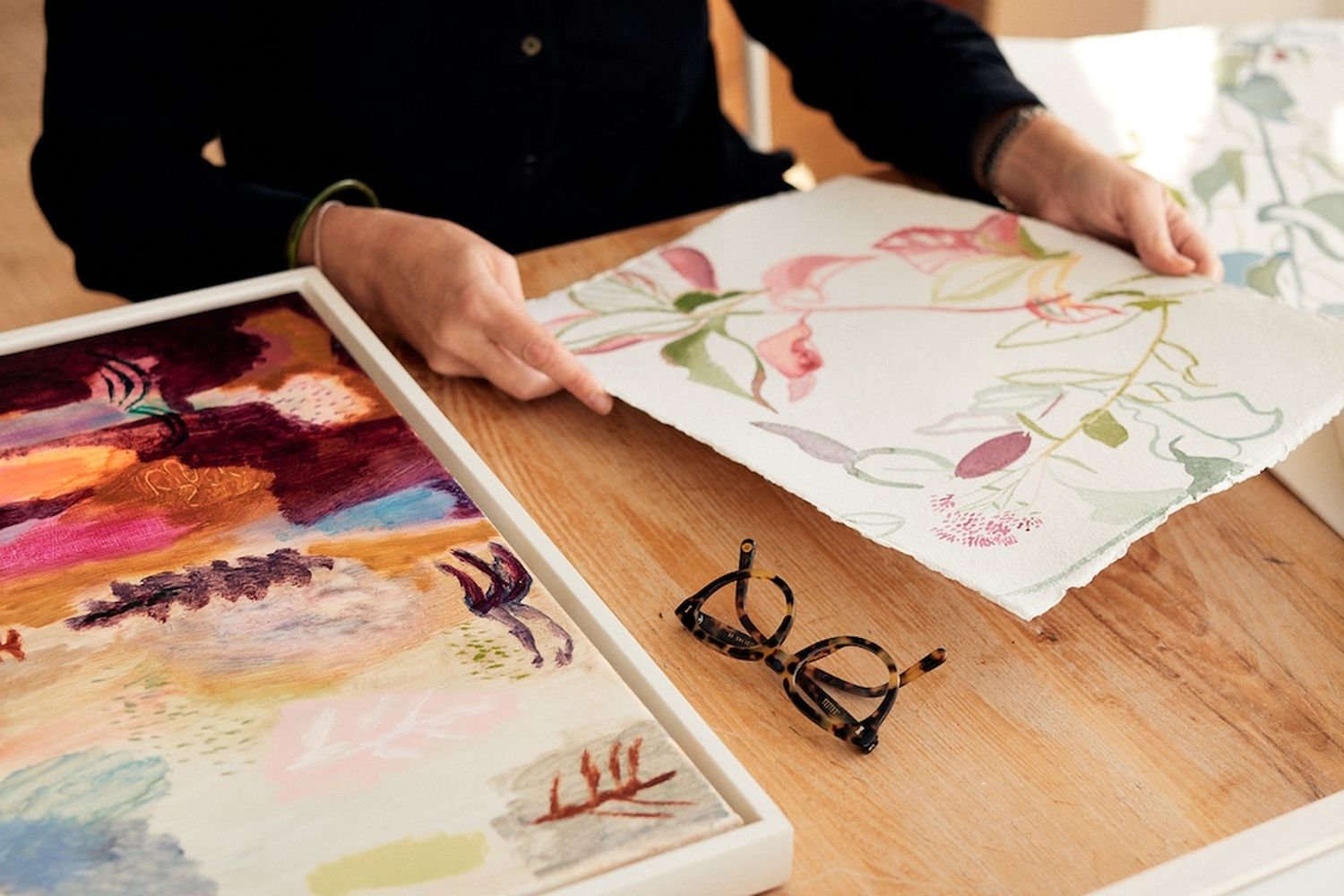 A person is working on a painting on a wooden table. They are holding a piece of paper and a pair of glasses. The painting is in a white frame and features a vibrant mix of colors. The person is wearing a black shirt. The table is also adorned with other paintings and a pair of glasses. [G]