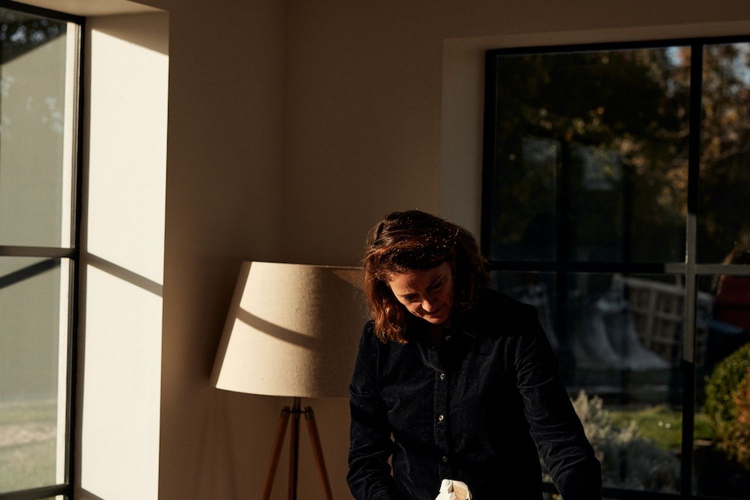 A woman in a black shirt is bending over a table, possibly sorting or arranging newspapers. The room has a window with a view of a garden outside. A lamp with a beige shade is on a table to the left of the woman. [G]