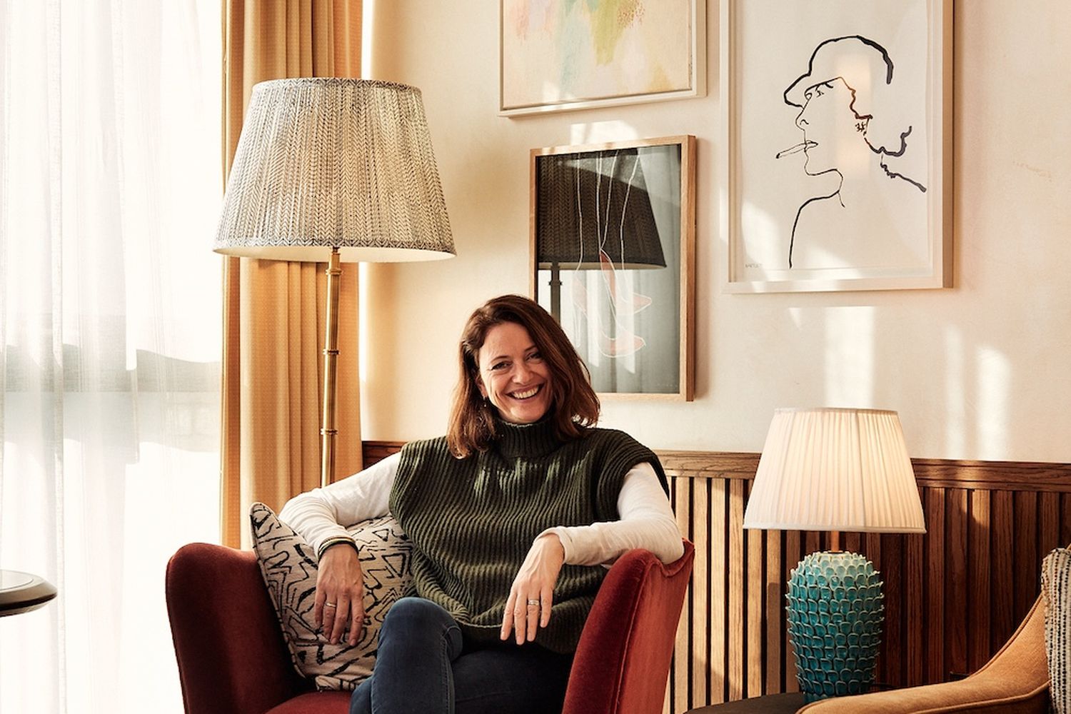 A woman in a green sweater and blue jeans, sitting on a red velvet chair, smiles at the camera. She is surrounded by a cozy living room with a window, a lamp, and a painting. [G]