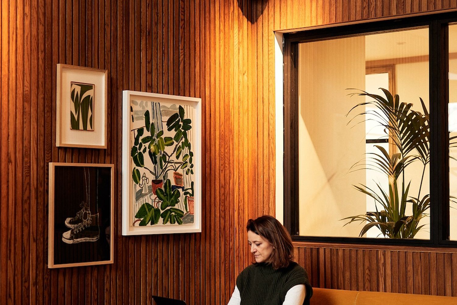 A woman is working at a laptop in a cozy cafe. The cafe has wooden walls and a large window. There are paintings on the wall, and a plant is in a corner. [G]
