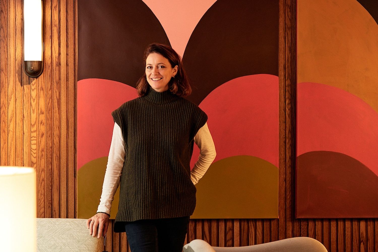 A woman in a green sweater stands in a modern living room with a large abstract painting on the wall. She is surrounded by a wooden table, a chair, and a lamp. [G]