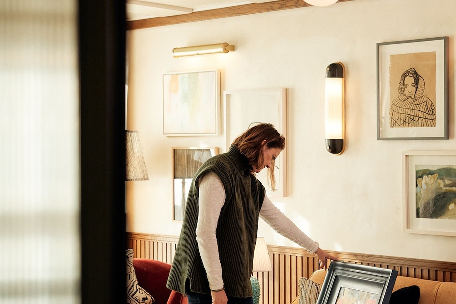 A man in a green vest is standing in a room with white walls and a wooden floor. He is reaching for a framed picture on a red couch. The room has several paintings on the walls and a lamp hanging from the ceiling. [G]