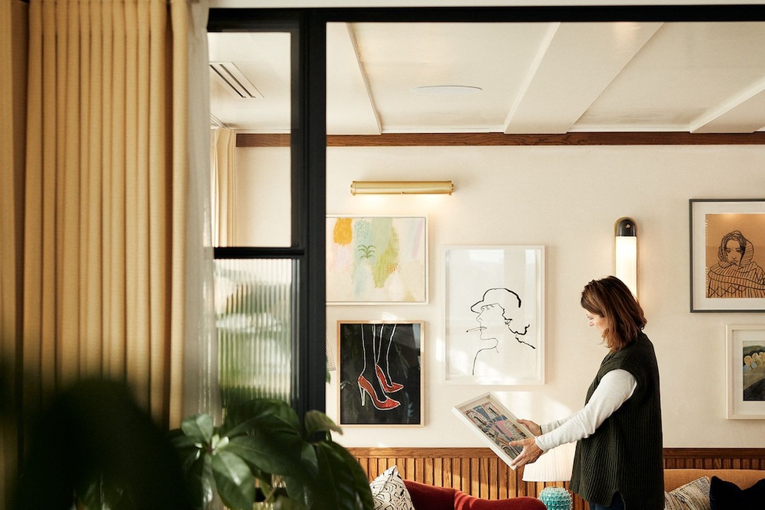 A woman in a green vest is reading a book in a cozy living room. The room has a large window with curtains, and there's a red sofa and a green plant in the corner. [G]