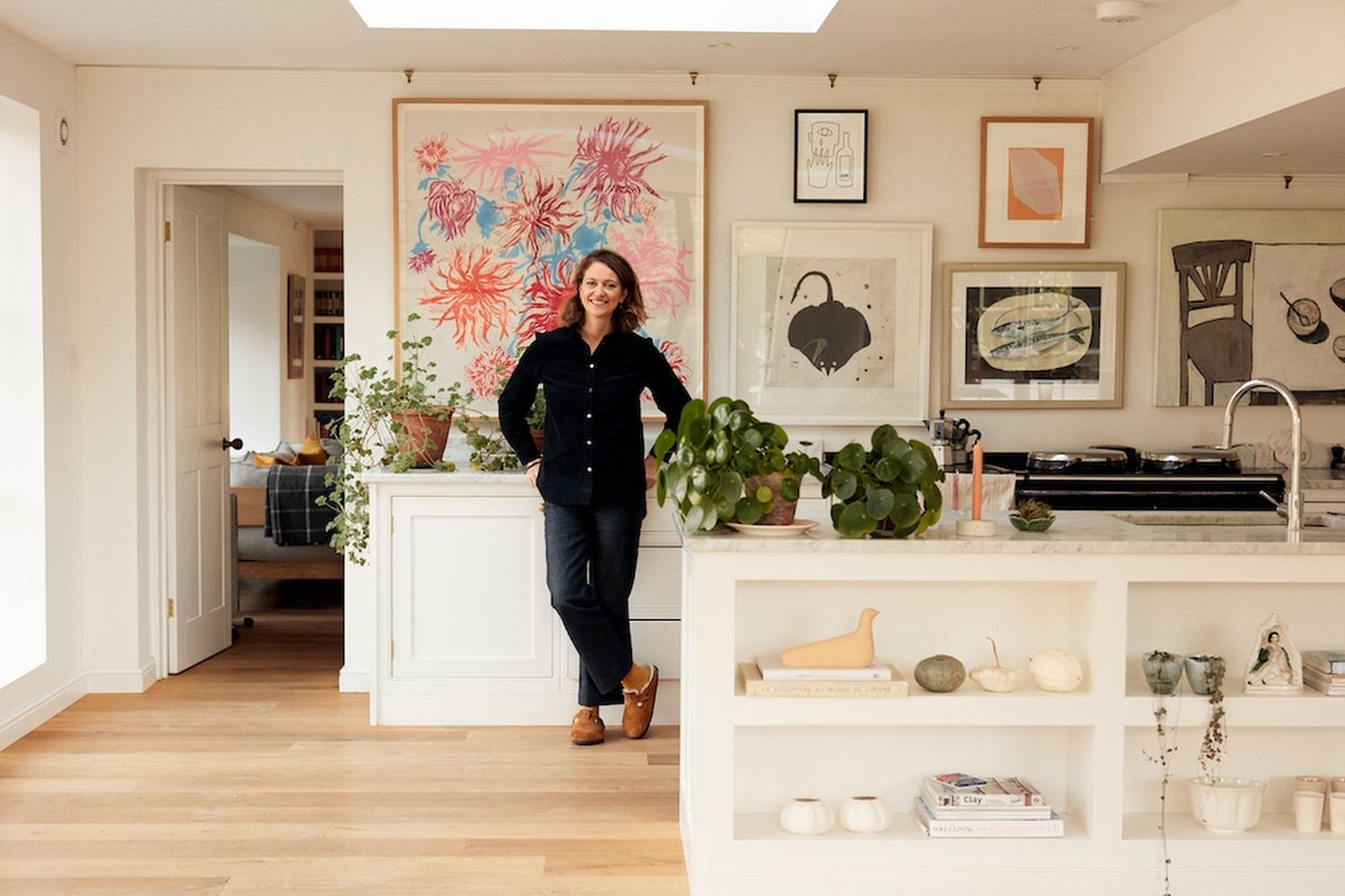 A woman in a black shirt and jeans stands in a cozy kitchen, surrounded by art and plants. [G]
