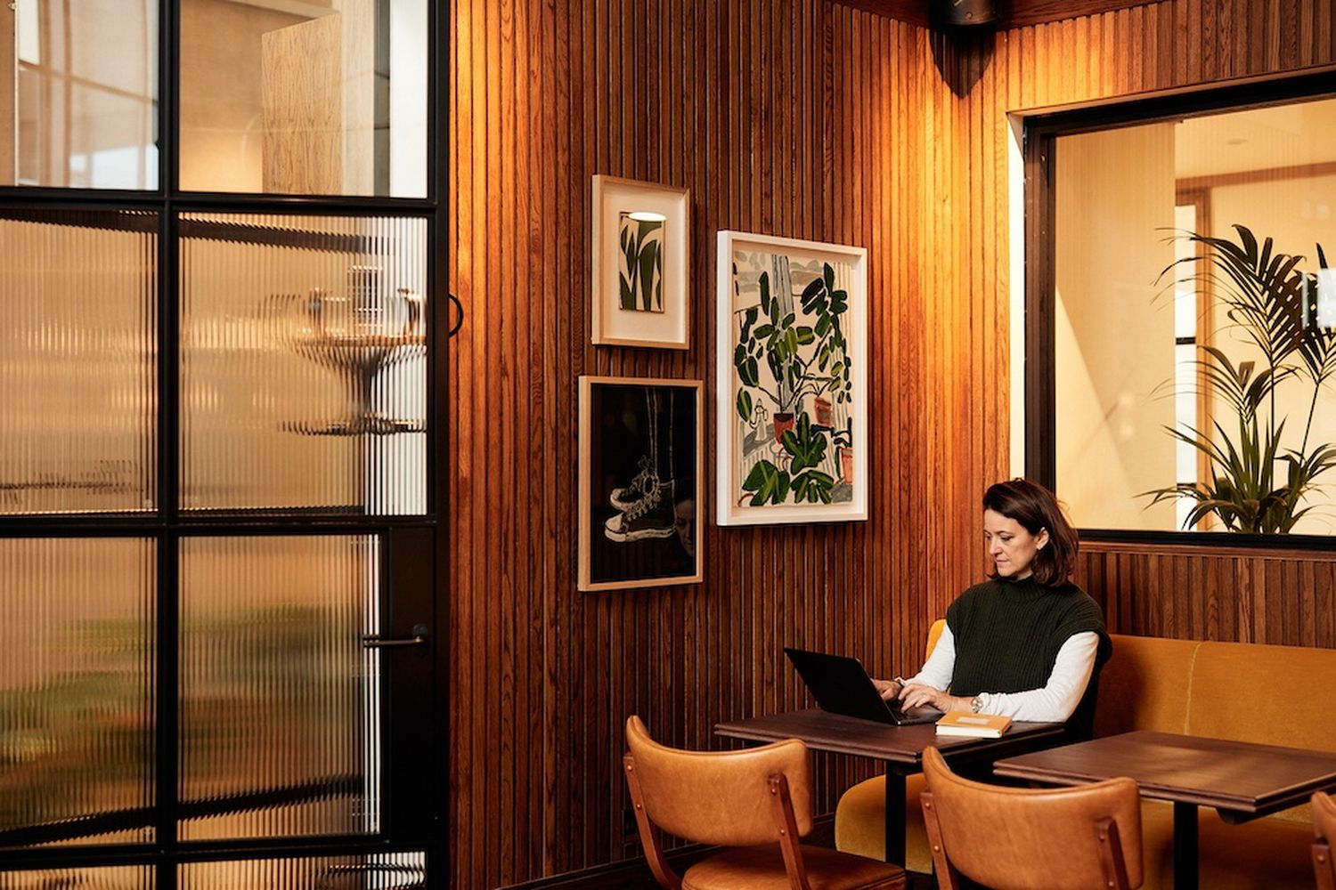 A woman is working at a table in a cozy room with wooden walls and a glass door. She is wearing a green sweater and has a laptop on her lap. There are several paintings on the wall, including a large one with a plant and a smaller one with a plant. The room also has a wooden door and a window. [G]